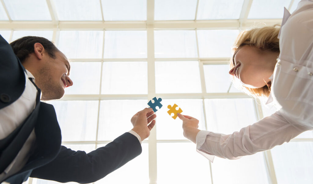 "Zwei Geschäftsleute fügen symbolisch Puzzleteile zusammen – ein Sinnbild für erfolgreiche Zusammenarbeit und wirtschaftliches Wachstum. In einem modernen Bürogebäude spiegelt das Bild die Vernetzung und strategische Partnerschaften in der Wirtschaft wider.