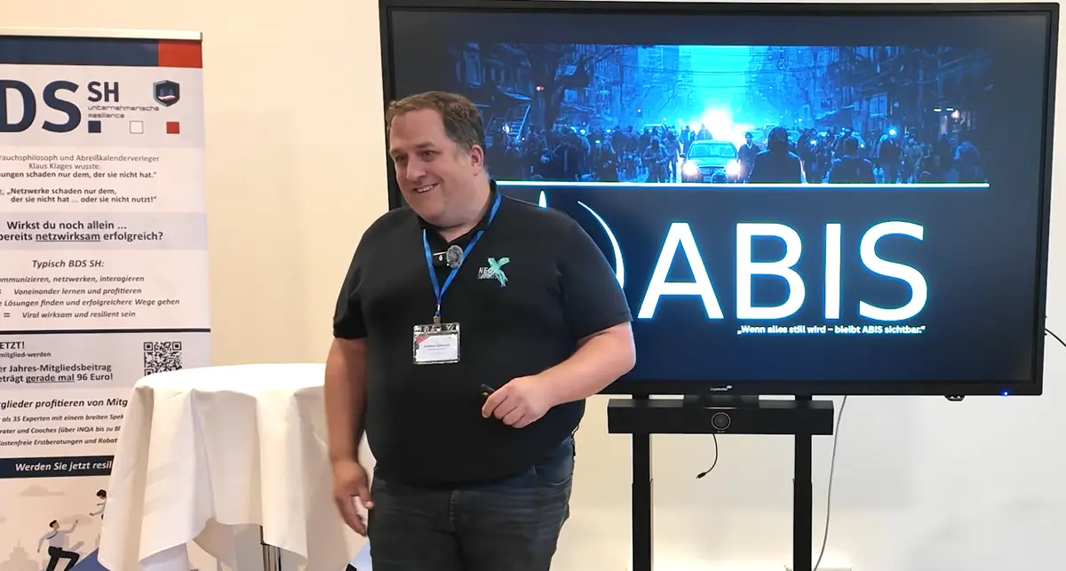 Ein Mann hält einen ABIS Vortrag vor einem Bildschirm mit der Aufschrift „ABIS“. Neben ihm steht ein Roll-up des BDS Schleswig-Holstein in einem Veranstaltungsraum.