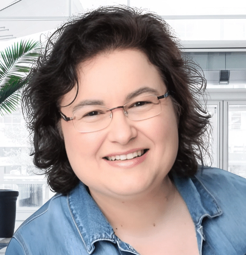 Simone Winkler lächelt freundlich in die Kamera. Sie trägt eine Brille und ein Jeanshemd. Im Hintergrund ist eine helle, moderne Büroumgebung mit großen Fenstern und Palmenpflanzen zu sehen.